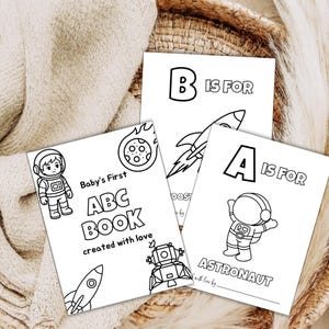 May include: Three white coloring pages featuring space-themed illustrations. One page reads Baby's First ABC BOOK created with love, another B IS FOR with a rocket, and the third A IS FOR ASTRONAUT with an astronaut illustration and a blank line.