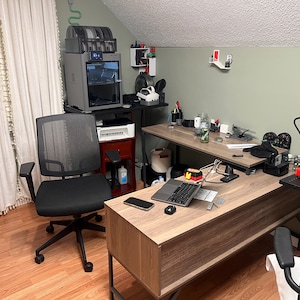 May include: A home office workspace featuring a black mesh ergonomic office chair, a wood-grain L-shaped desk with a laptop, a smartphone, and a 3D printer. A second 3D printer sits on a separate stand, alongside a white printer and various tools.