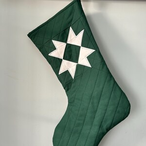 May include: A dark green quilted Christmas stocking with a white star design. The stocking has a diamond pattern and a hanging loop. A festive decoration for the holiday season.