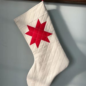 Peut inclure: Chaussette de Noël crème avec une étoile rouge appliquée. La chaussette est matelassée avec des coutures diagonales et est suspendue à un crochet en bois. Une boucle rouge est attachée en haut pour l'accrocher. L'étoile est d'un rouge vif.