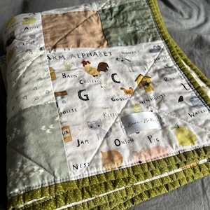 May include: A green and white patchwork quilt with a farm alphabet theme. The quilt features illustrations of farm animals, including a rooster, donkey, goose, kitten, pig, and a pear. The quilt is edged with a green ruffled trim and has a white label that reads "Bear Belle".