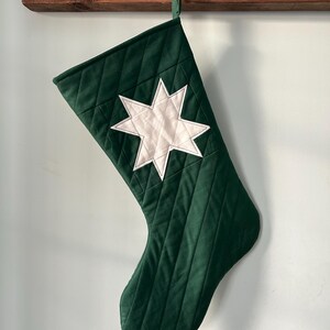 May include: A green quilted Christmas stocking with a large, off-white star applique. The stocking is suspended from a wooden rack with black hooks. The fabric has a diamond pattern. The stocking is a festive decoration.