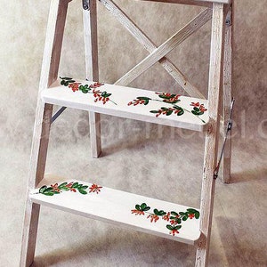 Puede incluir: Un taburete de madera blanco con tres escalones. Los escalones y la parte superior están decorados con un diseño floral de hojas verdes y bayas rojas. El taburete tiene un acabado envejecido y es adecuado para la decoración del hogar.