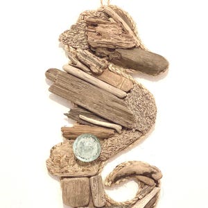 Puede incluir: Un adorno de pared con forma de caballito de mar hecho con madera flotante, cuerda y una canica de cristal. El caballito de mar está compuesto por piezas de madera desgastada de diferentes tamaños y formas, con cuerda delineando la forma.