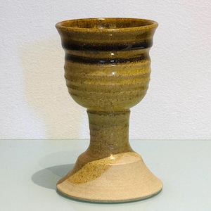 May include: A handmade ceramic goblet with a wide, textured rim and a pedestal base. The goblet features a blend of earthy tones, including shades of brown, tan, and olive green. The design has a rustic, artisanal aesthetic.