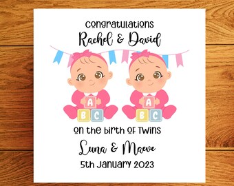 May include: A white card with a congratulations message for Rachel & David on the birth of twins Luna & Maeve on January 5th, 2023. The card features an illustration of two baby girls with pink bows and pink outfits holding colorful blocks.