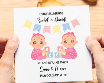 May include: A pink and white card with a congratulations message for Rachel & David on the birth of twins Luna & Maeve on October 14th, 2022. The card features a cartoon illustration of two babies playing with blocks.