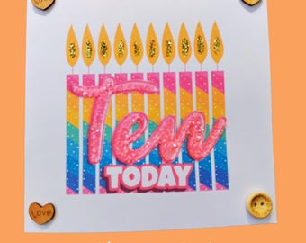 May include: A white card with a pink and yellow glittery "Ten Today" design. The card has a rainbow striped background with yellow candles at the top. The card is decorated with small wooden hearts that say "Love" and a wooden button.