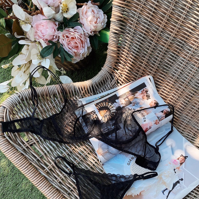 May include: Black lace lingerie set, including a bra and a thong, displayed on a wicker chair with a magazine and a bouquet of pink and white flowers. The bra has delicate straps and intricate lace detailing.