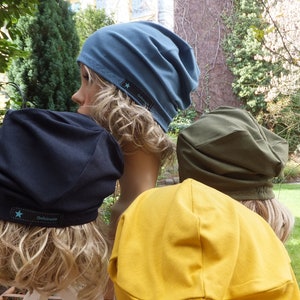 Könnte beinhalten: Eine Auswahl an Slouch-Beanies in Marineblau, Türkis, Olivgrün und Senfgelb. Jeder Hut hat ein kleines schwarzes Etikett mit einem Stern und dem Wort "Defiance". Die Hüte werden auf Schaufensterpuppenköpfen präsentiert.