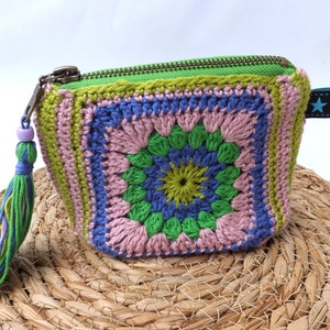 Könnte beinhalten: Bunte, gehäkelte quadratische Tasche mit Reißverschluss. Das Täschchen hat ein Granny-Square-Design in Grün, Blau und Rosa. Ein Quastenanhänger und ein kleines Stern-Etikett ergänzen die Dekoration. Ca. 10 cm breit.