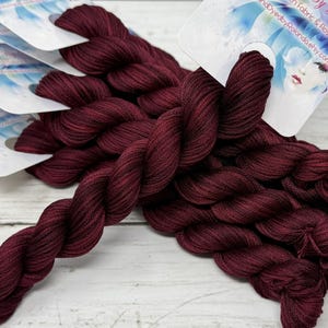 May include: Bundles of burgundy embroidery floss with attached tags. The floss is tightly wound, highlighting its texture and colour. The tags feature a watercolour design and text. The bundles are arranged on a light-coloured wooden surface.