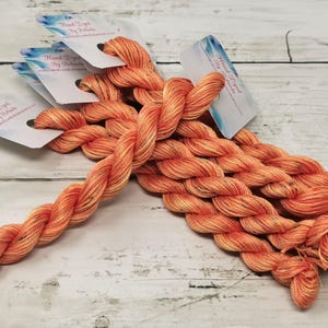 Puede incluir: Primer plano de un conjunto de hilo de bordar teñido a mano de color naranja. El hilo está retorcido y tiene un brillo sutil. El hilo está etiquetado como "Teñido a mano por Rebecca".