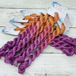 May include: A collection of hand-dyed embroidery floss skeins featuring a vibrant gradient transition from deep purple at the bottom to a warm golden orange at the top. Each twisted thread bundle is secured with a white paper tag labeled Hand Dyed.