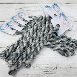 May include: Several skeins of hand-dyed embroidery floss in shades of gray and blue. Each skein is twisted and has a tag with the text "Hand Dyed by Rebecca". The floss is arranged on a white wooden surface.
