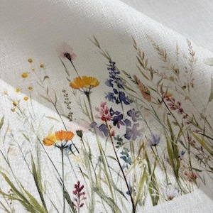 May include: A white fabric featuring a printed pattern of watercolour wildflowers and grasses. The design includes yellow, orange, purple, and pink blossoms with green stems and stalks, arranged in a natural, scattered meadow-like composition.