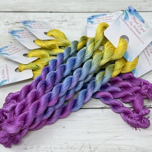 Könnte beinhalten: Mehrere Stränge handgefärbtes Stickgarn mit einem bunten Farbverlauf, der von hellem Gelb oben über Hellblau in der Mitte bis zu tiefem Violett unten reicht, wobei jeder Strang an einem mit einem Markennamen versehenen Papieretikett befestigt ist.