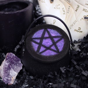 May include: A small black cauldron with a pentagram design on the front. The pentagram is purple with a glittery finish. The cauldron is sitting on black shredded paper.
