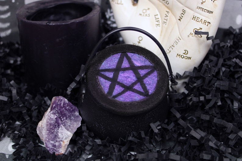 May include: A small black cauldron with a pentagram design on the front. The pentagram is purple with a glittery finish. The cauldron is sitting on black shredded paper.