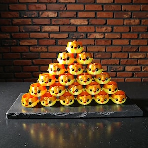 Peut inclure: Une pyramide de récipients en forme de poussin jaunes et oranges. Chaque récipient a un visage souriant avec des yeux noirs et un bec. Les récipients sont empilés sur une surface rectangulaire gris foncé devant un mur de briques.