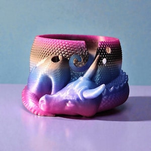Könnte beinhalten: Ein 3D-gedruckter Drachen-Blumentopf in Rosa-, Blau- und Lilatönen. Der Drache ist um den Topf gewickelt, sein Kopf ruht an der Seite. Der Topf hat eine strukturierte Oberfläche und eine runde Öffnung.