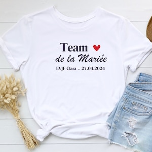 Peut inclure: T-shirt blanc avec le texte "Team de la Mariée" en bleu marine avec un cœur rouge. Le texte "EVJF Clara - 27.04.2024" est également imprimé en bleu marine.
