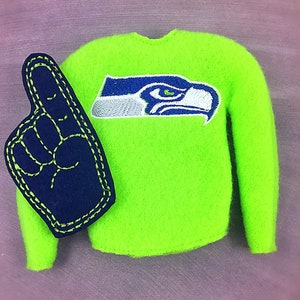 May include: A neon green felt sweatshirt with the Seattle Seahawks logo embroidered in blue and white. A navy blue foam finger with yellow stitching is next to the sweatshirt. The items are on a purple background.