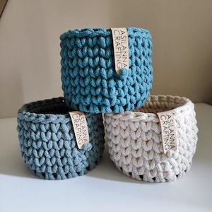 Può includere: Tre cestini all'uncinetto fatti a mano nei toni del blu e del beige. Ogni cestino ha una forma cilindrica con un motivo a punto strutturato. Una piccola etichetta con la scritta "ASILANNA CRAFTING" è attaccata a ciascun cestino.