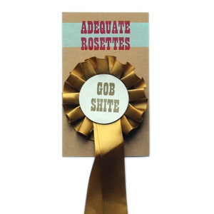 Può includere: Una rosetta dorata con un cerchio bianco al centro che dice "Gob Shite". La rosetta è attaccata a una carta marrone che dice "Adequate Rosettes".