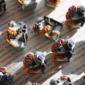 May include: Miniature polymer clay sculptures of stacked books adorned with roses and dragon accents. The books are in shades of blue, green, and brown, with pink, blue, and orange flowers. The sculptures are on a book page.