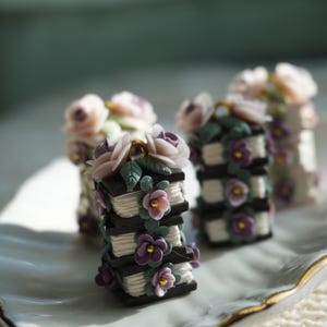 May include: Miniature stacked book sculptures with dark brown covers and white pages, adorned with pastel pink and purple flowers and green leaves. The books are arranged on a white plate with a gold rim, creating a whimsical, floral theme.