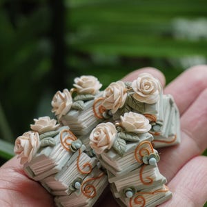 May include: Miniature stacks of books with cream-colored roses and green leaves. The books are light blue-green with orange and blue accents. The floral arrangement is held in a hand.