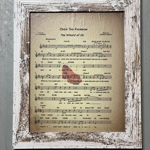 May include: A vintage sheet music print of "Over the Rainbow" from the movie "The Wizard of Oz". The sheet music is framed in a white distressed wood frame. The lyrics and musical notes are printed on a cream-colored paper.