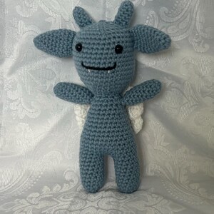 Puede incluir: Una criatura de ganchillo azul hecha a mano con orejas grandes, alas peque&ntilde;as y una boca sonriente. El peluche tiene ojos de bot&oacute;n negros y est&aacute; de pie. Las alas son blancas. El fondo es una tela blanca estampada.