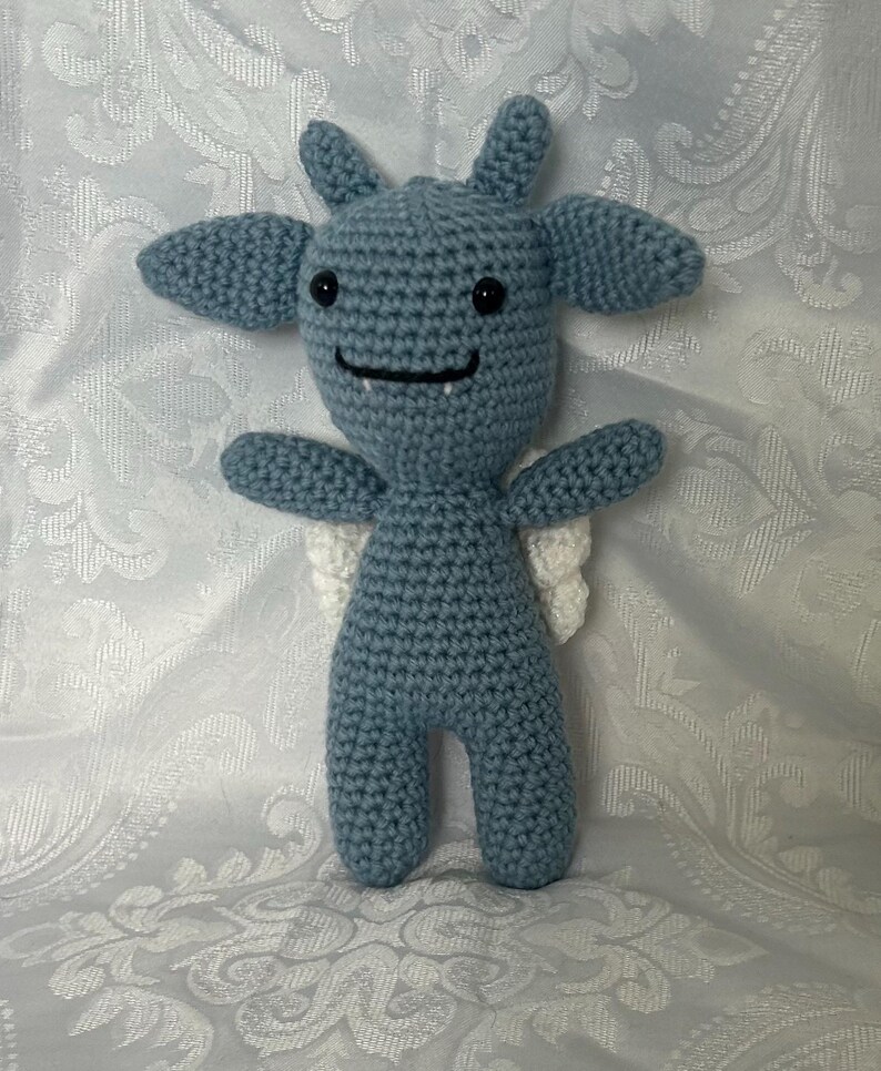 Puede incluir: Una criatura de ganchillo azul hecha a mano con orejas grandes, alas peque&ntilde;as y una boca sonriente. El peluche tiene ojos de bot&oacute;n negros y est&aacute; de pie. Las alas son blancas. El fondo es una tela blanca estampada.