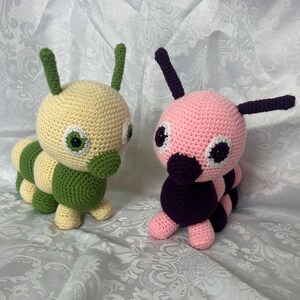 May include: Two crocheted caterpillar plush toys. One is yellow and green, the other is pink and purple. Both have large, round eyes and small antennae. The toys are handmade and appear soft and cuddly.
