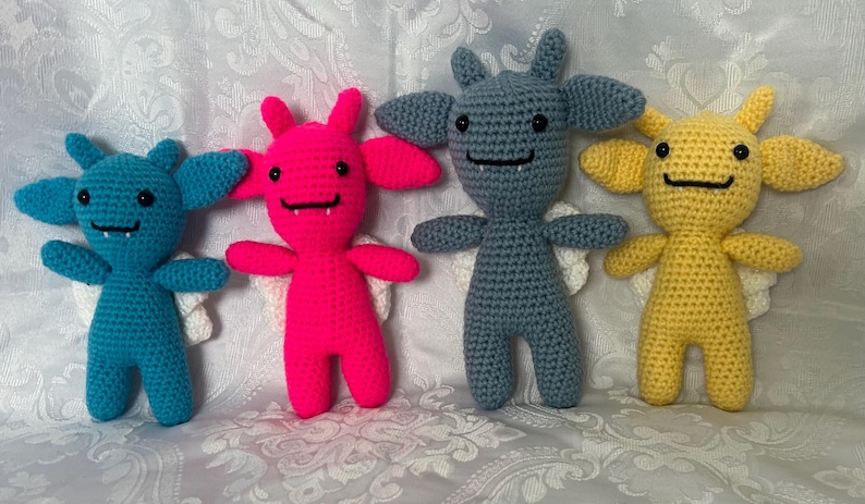 Puede incluir: Cuatro mu&ntilde;ecos de monstruo hechos a mano con ganchillo en azul, rosa, gris y amarillo. Cada mu&ntilde;eco tiene orejas grandes, alas peque&ntilde;as y una cara sonriente. Los mu&ntilde;ecos miden unos 15 cm de alto.