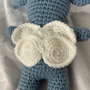Puede incluir: Mu&ntilde;eca de ganchillo azul claro con alas blancas. La mu&ntilde;eca est&aacute; hecha a mano, con una superficie texturizada creada por los puntos de ganchillo. Las alas son de hilo blanco y est&aacute;n unidas a la espalda de la mu&ntilde;eca.