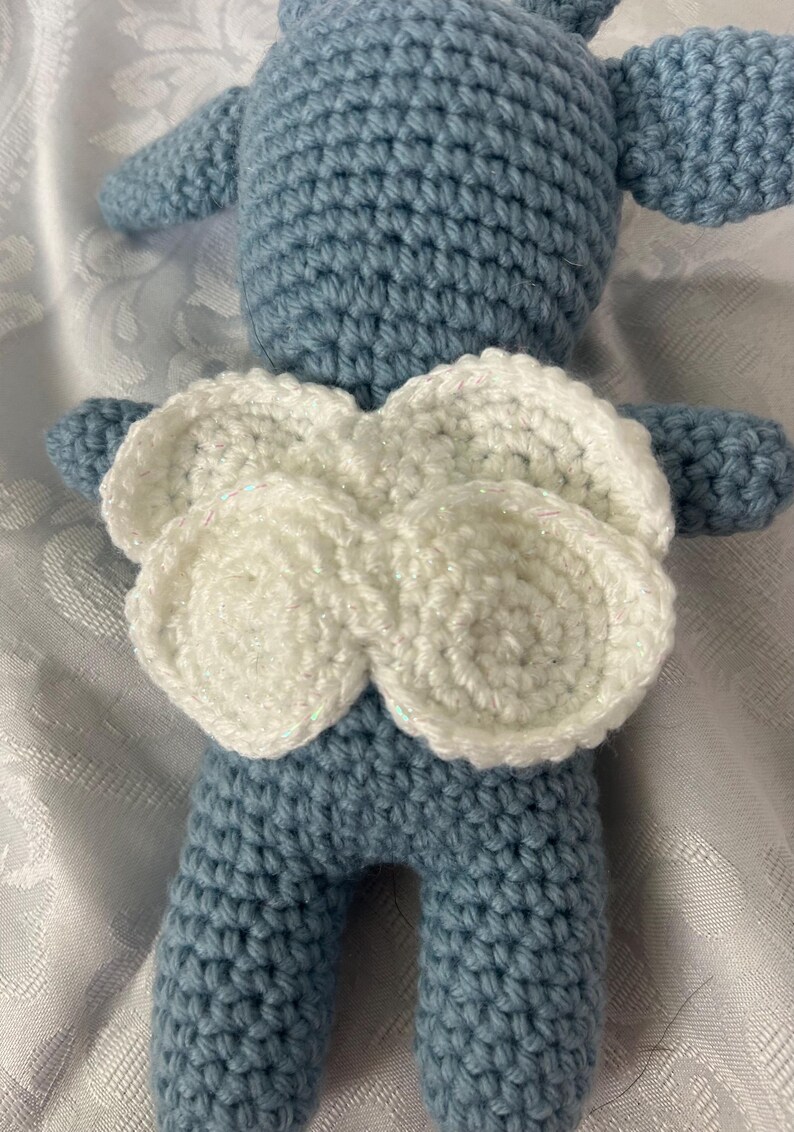 Puede incluir: Mu&ntilde;eca de ganchillo azul claro con alas blancas. La mu&ntilde;eca est&aacute; hecha a mano, con una superficie texturizada creada por los puntos de ganchillo. Las alas son de hilo blanco y est&aacute;n unidas a la espalda de la mu&ntilde;eca.