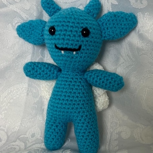 Puede incluir: Una criatura de ganchillo turquesa hecha a mano con orejas grandes, peque&ntilde;as alas blancas y una cara sonriente. El peluche tiene ojos de bot&oacute;n negros y una boca peque&ntilde;a con un solo diente. Los brazos y las piernas tambi&eacute;n est&aacute;n hechos a ganchillo.