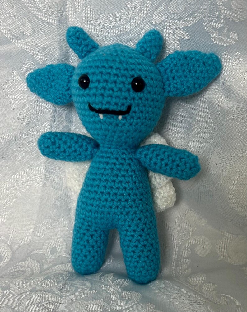 Puede incluir: Una criatura de ganchillo turquesa hecha a mano con orejas grandes, peque&ntilde;as alas blancas y una cara sonriente. El peluche tiene ojos de bot&oacute;n negros y una boca peque&ntilde;a con un solo diente. Los brazos y las piernas tambi&eacute;n est&aacute;n hechos a ganchillo.
