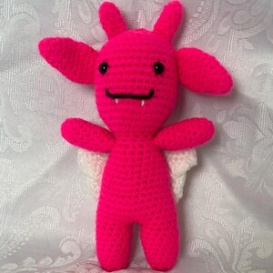 Puede incluir: Mu&ntilde;eca de ganchillo rosa brillante hecha a mano, con ojos negros, una sonrisa negra y dos peque&ntilde;os colmillos. La mu&ntilde;eca tiene orejas grandes, cuernos peque&ntilde;os y alas blancas. La mu&ntilde;eca est&aacute; de pie.