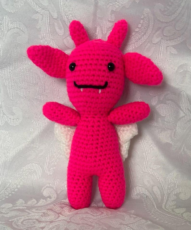 Puede incluir: Mu&ntilde;eca de ganchillo rosa brillante hecha a mano, con ojos negros, una sonrisa negra y dos peque&ntilde;os colmillos. La mu&ntilde;eca tiene orejas grandes, cuernos peque&ntilde;os y alas blancas. La mu&ntilde;eca est&aacute; de pie.