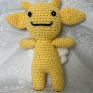 Puede incluir: Una criatura amarilla tejida a mano con ojos negros, una boca sonriente y peque&ntilde;os colmillos. Tiene dos cuernos peque&ntilde;os, dos ap&eacute;ndices grandes en forma de oreja, brazos peque&ntilde;os y alas blancas. El peluche est&aacute; de pie.