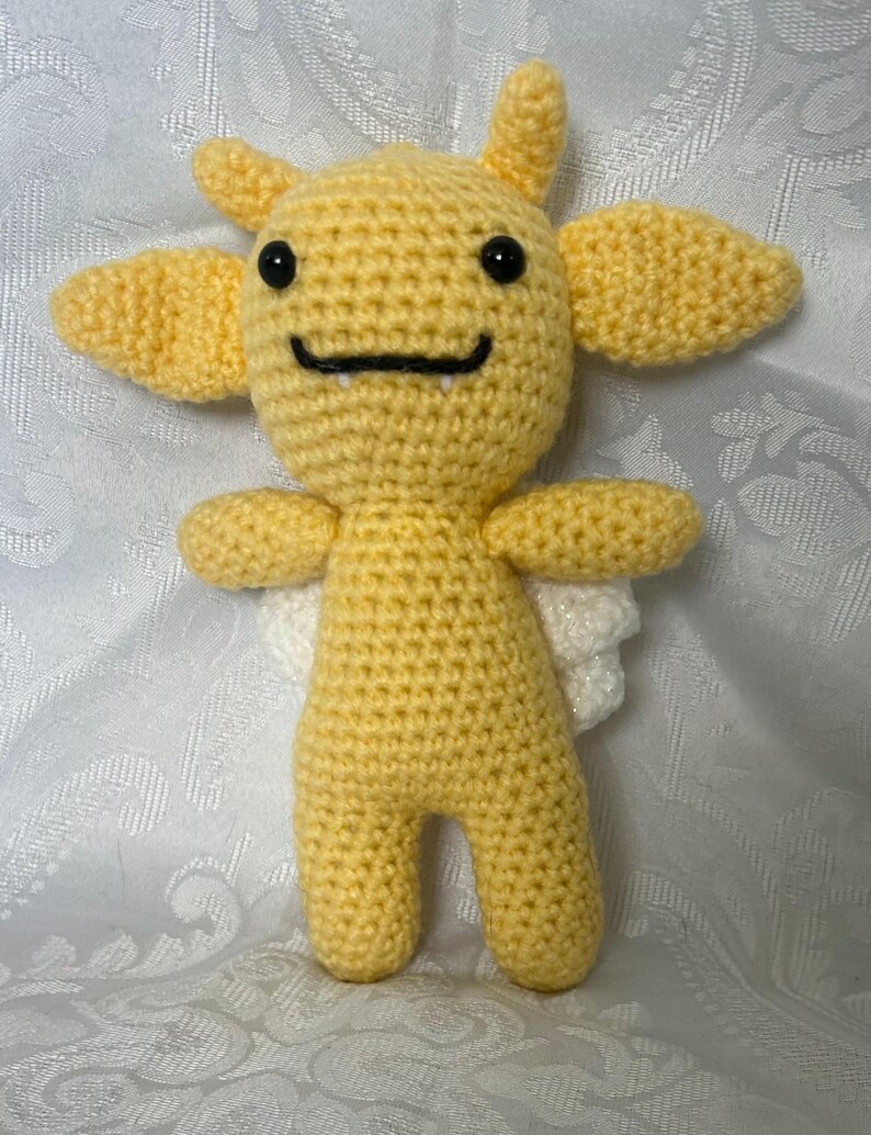 Puede incluir: Una criatura amarilla tejida a mano con ojos negros, una boca sonriente y peque&ntilde;os colmillos. Tiene dos cuernos peque&ntilde;os, dos ap&eacute;ndices grandes en forma de oreja, brazos peque&ntilde;os y alas blancas. El peluche est&aacute; de pie.