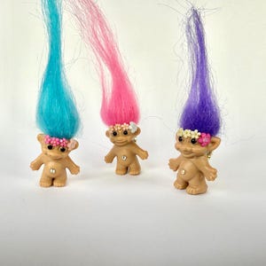 May include: Three vintage troll dolls with colorful hair in blue, pink, and purple. Each doll has a flower crown and a jewel on its belly. The dolls are standing on a white surface.
