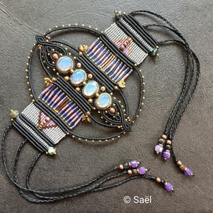 Op de afbeelding: Een handgemaakte macramé ketting met een centraal ontwerp met vier ovale, opalescente stenen. De ketting bevat zwarte, grijze, roze en gouden accenten, met kralen details en lange zwarte koorden. Het ontwerp bevat de tekst "© Saël".