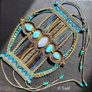 Puede incluir: Un collar de macramé hecho a mano con piedras preciosas turquesas y cuentas iridiscentes. El intrincado diseño incorpora detalles dorados y una piedra lunar central. El collar tiene una forma geométrica única y se muestra sobre una superficie oscura.