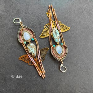 May include: Two long dangle earrings featuring macrame cord in brown and pink, accented with teardrop-shaped moonstone and turquoise gemstones. Each earring incorporates a gold-toned metal moth wing cutout and small beads. Text reads Saël.