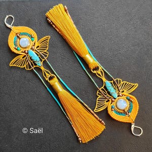 Peut inclure: Une paire de longues boucles d'oreilles avec des pompons en fil jaune moutarde, des éléments métalliques dorés en forme de papillon, des cabochons irisés ronds et des pierres allongées turquoise. Le texte Saël est visible en bas à gauche.